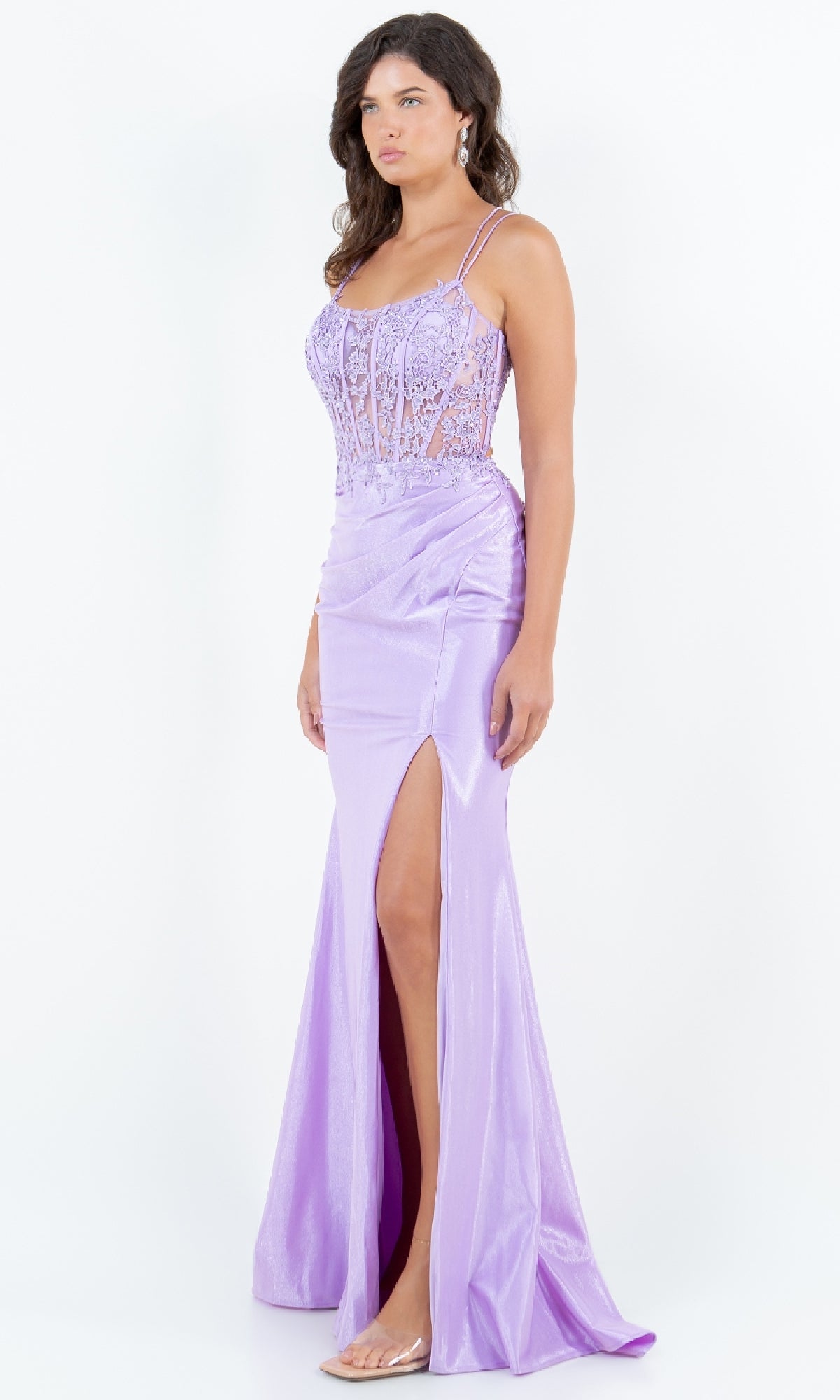 A woman models the Lace-Up Lilac Prom Gown: Cinderella Couture 8185J—featuring a floor-length ruched skirt, sheer lace bodice, thin straps, lace-up back, and high slit—against a white background.